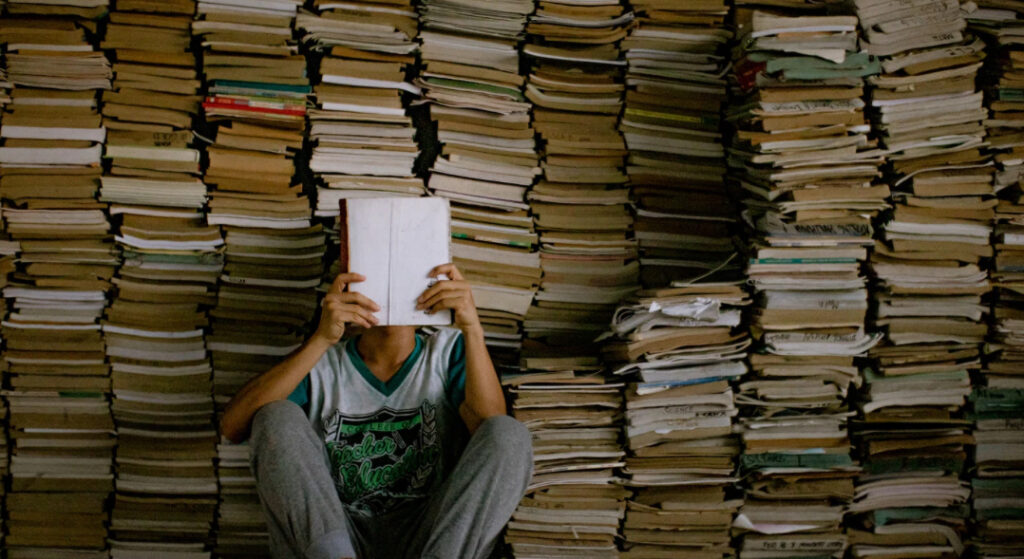 Imagen de un hombre leyendo con una pila de libros a su espalda (Canva)